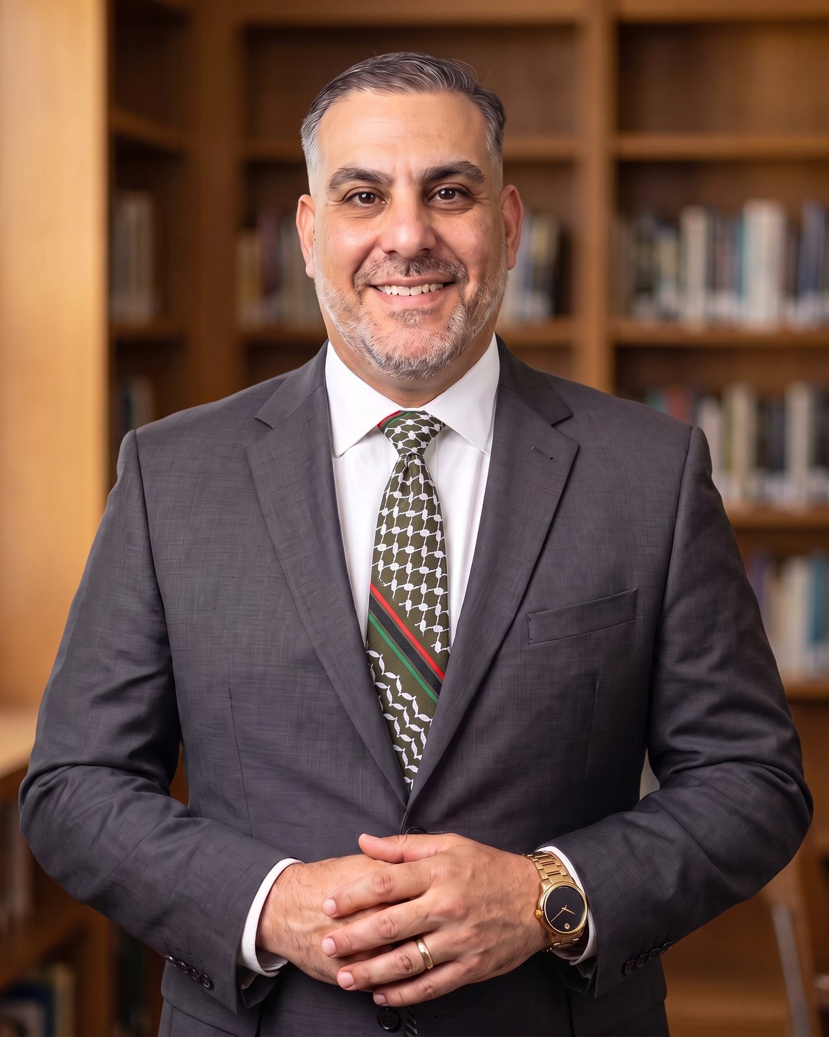 Dr. Kareem Tannous, portrait in a charcoal-gray suit and the Zaytoun necktie, olive green under a white keffiyeh net, standing before a private-library bookshelf, gold band and Movado watch visible.